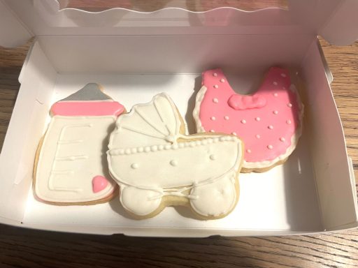 Palette gateaux biscuits baby shower Trois biscuits en forme de bouteille, landau et robe, décorés en blanc et rose.