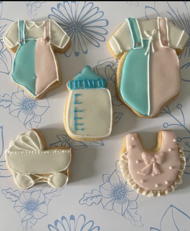 Gateaux biscuits baby shower naissance Des biscuits en format de vêtements de bébé, biberon et landau, décorés en pastel.